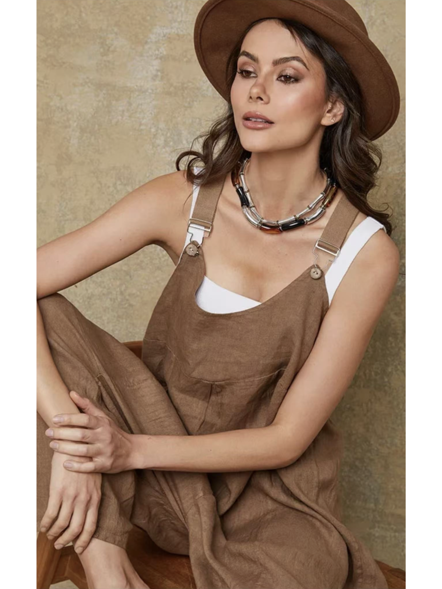Model in a camel 100% linen jumpsuit with a relaxed fit, adjustable buckle straps, and button details at the ankles, over a white tank top.