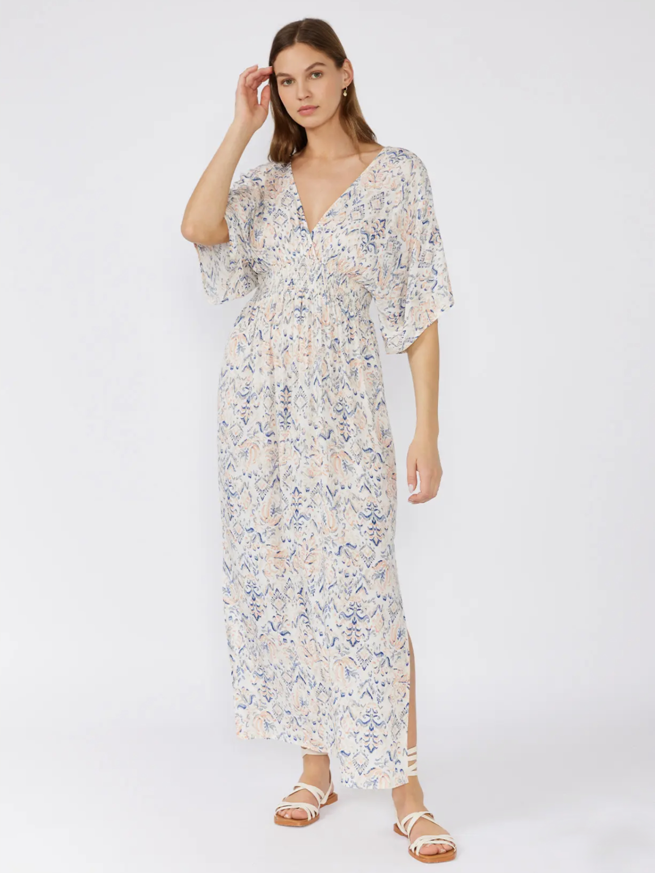 Woman in a white bohemian maxi dress with a blue and peach paisley print, kimono sleeves, and a smocked waist, standing against a white background.