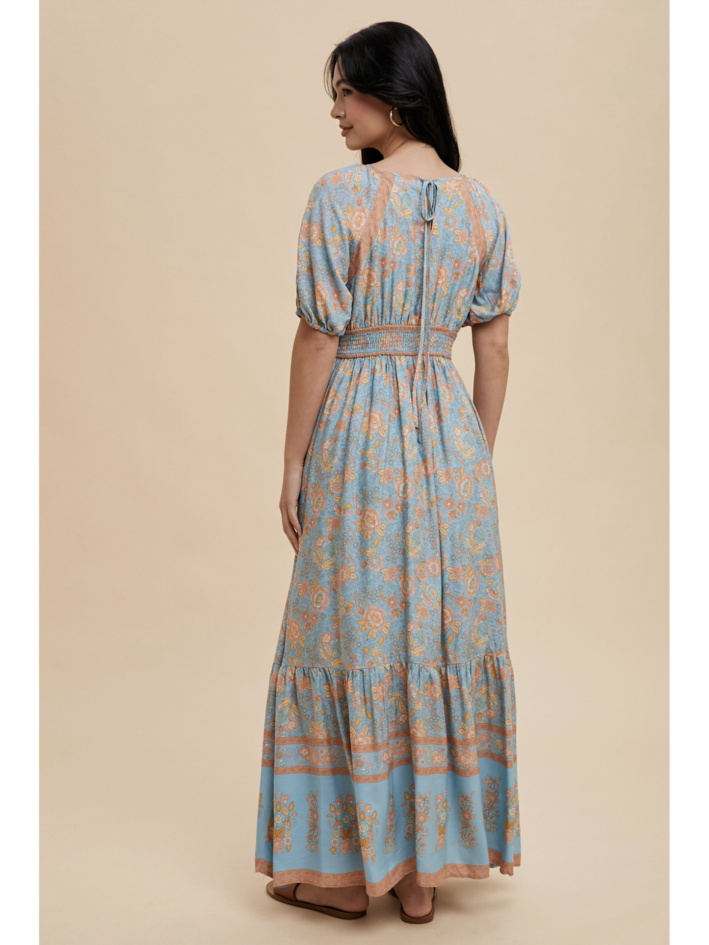 A woman wearing a dusty blue maxi dress with white botanical border prints, lace trim, standing against a soft-focus background.