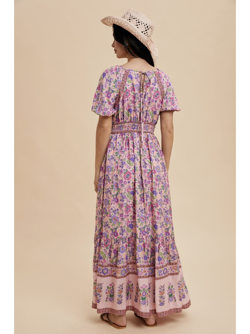 A woman wearing a pink fuschia maxi dress with white botanical border prints, lace trim, standing against a soft-focus background.