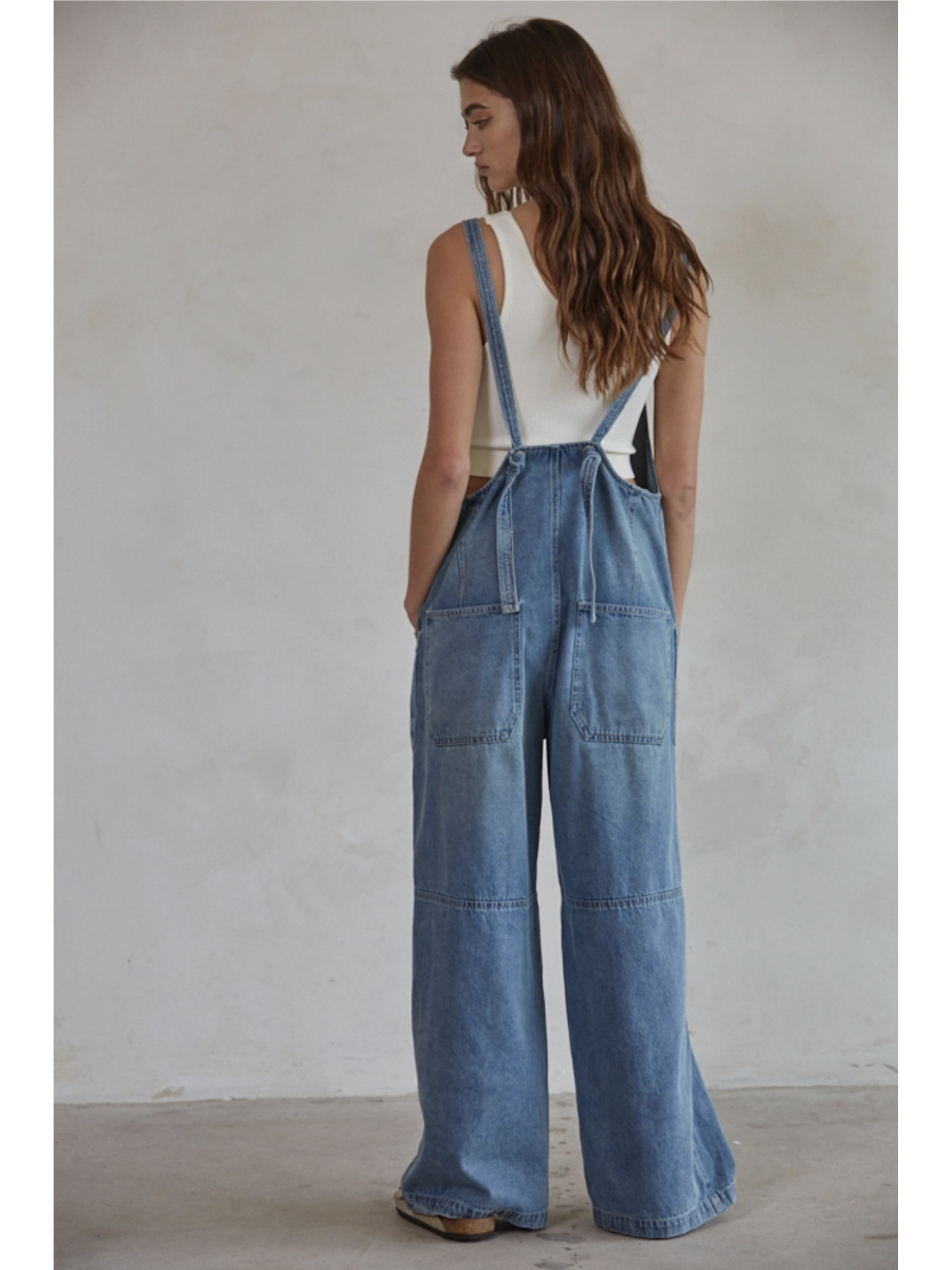Model in blue denim wide-leg overalls with a square neck and button-down front, styled over a white t-shirt against a plain background.