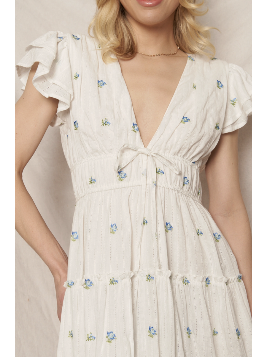 Woman in a white tiered maxi dress with a blue and peach floral pattern, featuring flutter sleeves, standing against a clean white background.