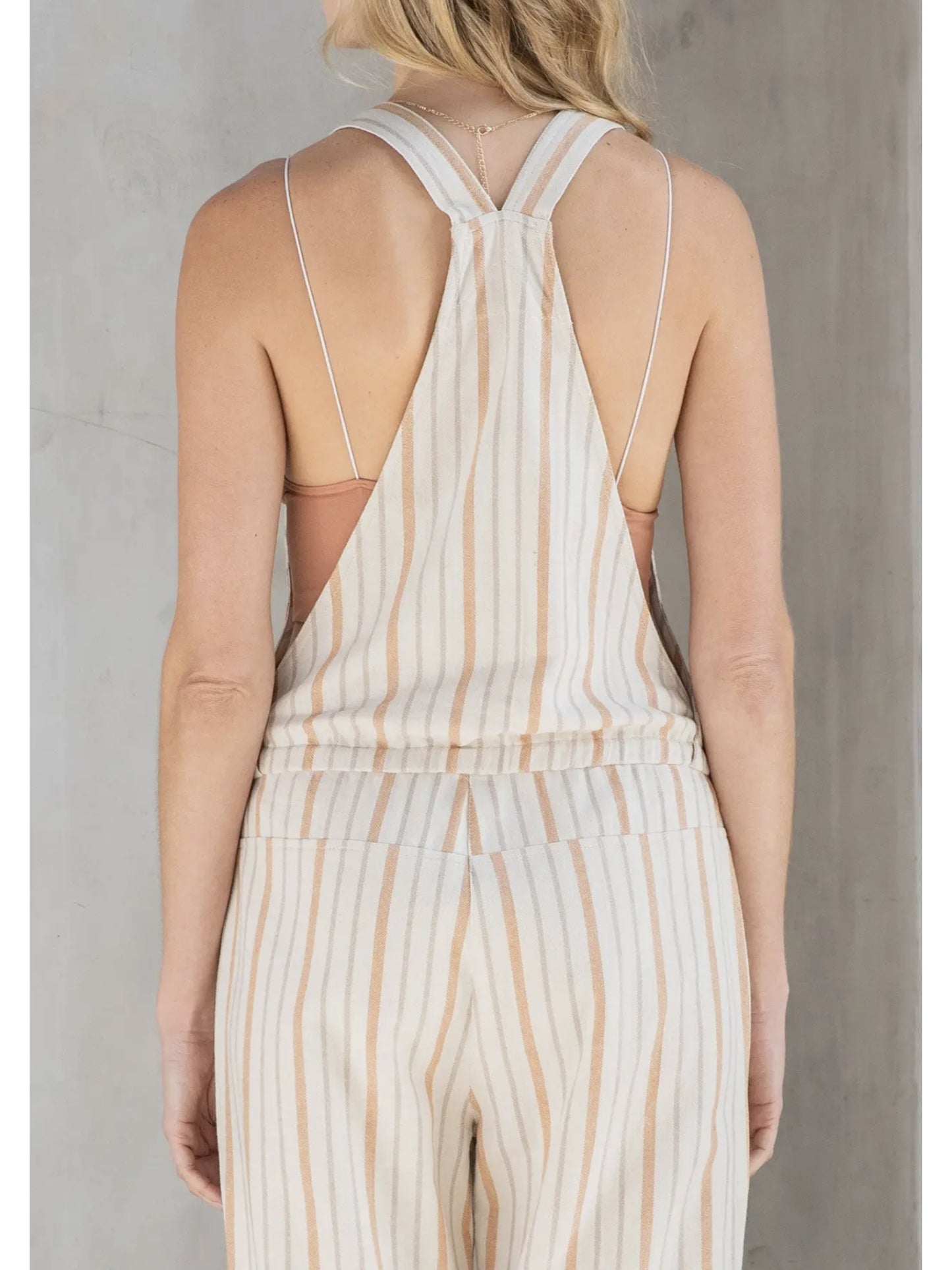 Woman in cream and tan striped linen-blend overalls with a drawstring waist and front zipper pocket, standing against a grey textured wall.