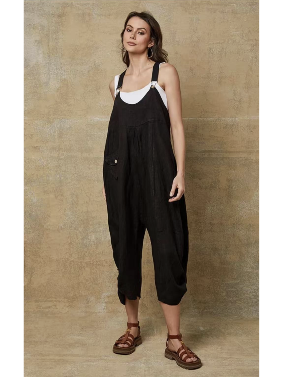 Model in a black 100% linen jumpsuit with a relaxed fit, adjustable buckle straps, and button details at the ankles, over a white tank top.