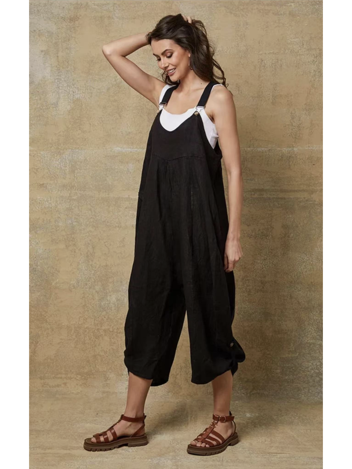 Model in a black 100% linen jumpsuit with a relaxed fit, adjustable buckle straps, and button details at the ankles, over a white tank top.