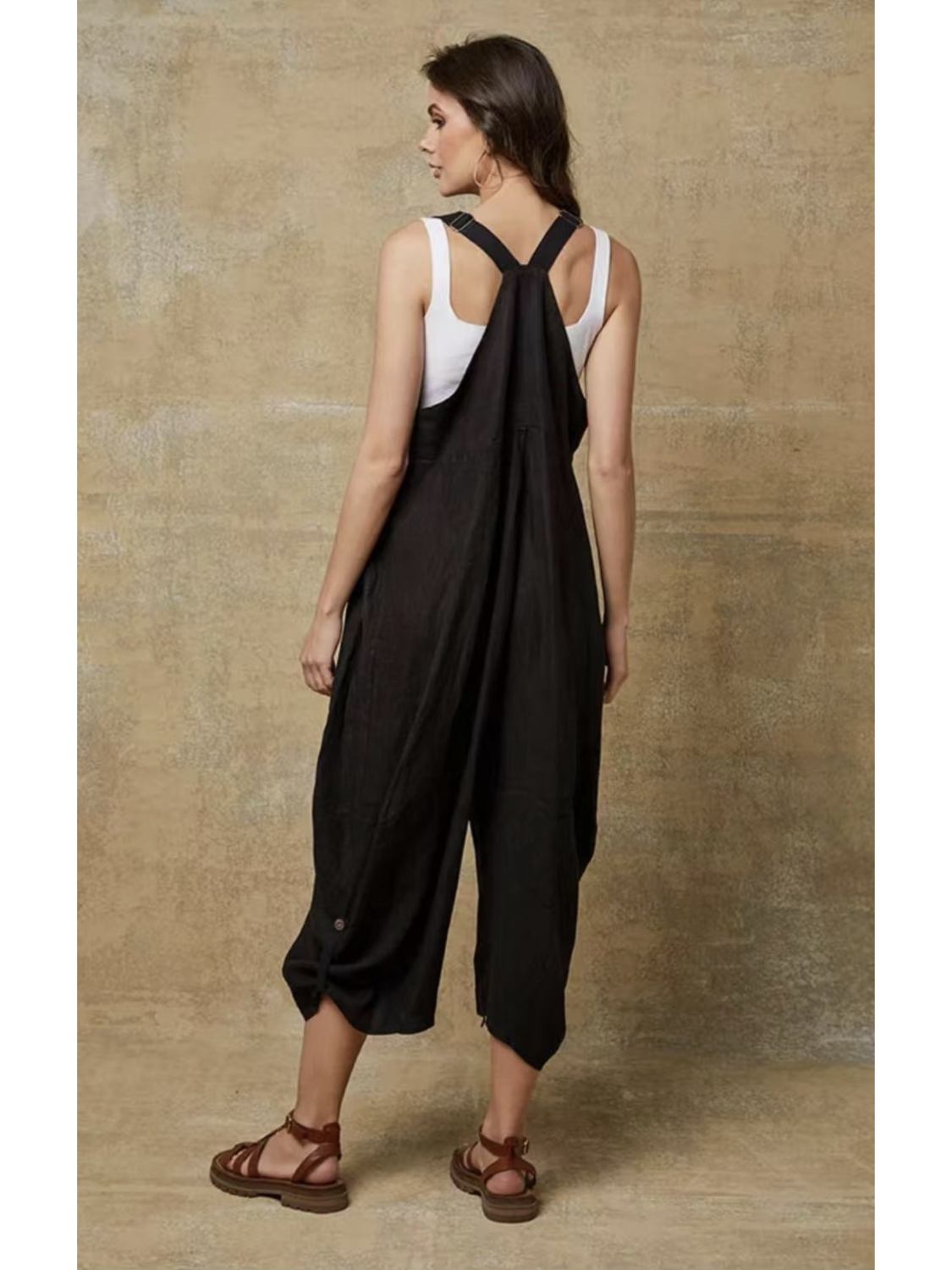 Model in a black 100% linen jumpsuit with a relaxed fit, adjustable buckle straps, and button details at the ankles, over a white tank top.