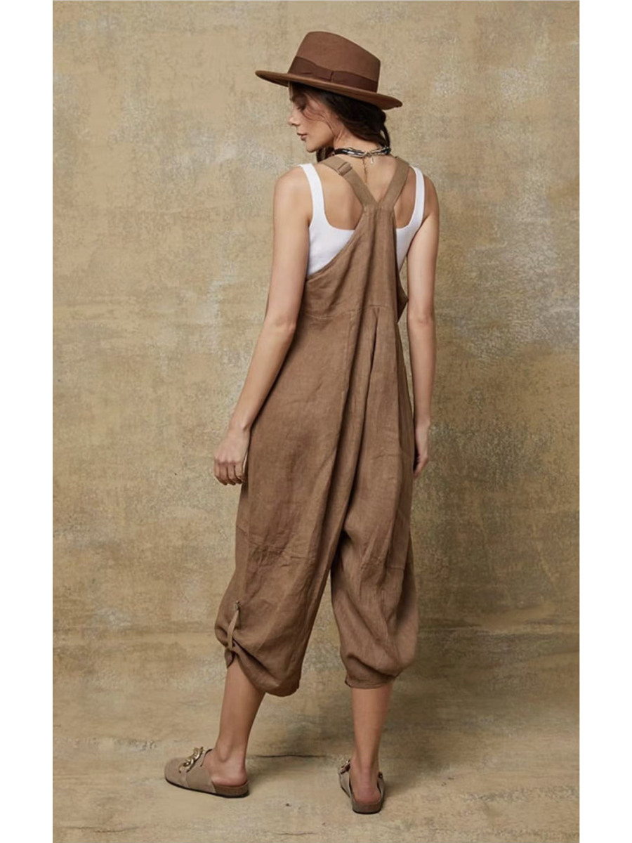 Model in a camel 100% linen jumpsuit with a relaxed fit, adjustable buckle straps, and button details at the ankles, over a white tank top.
