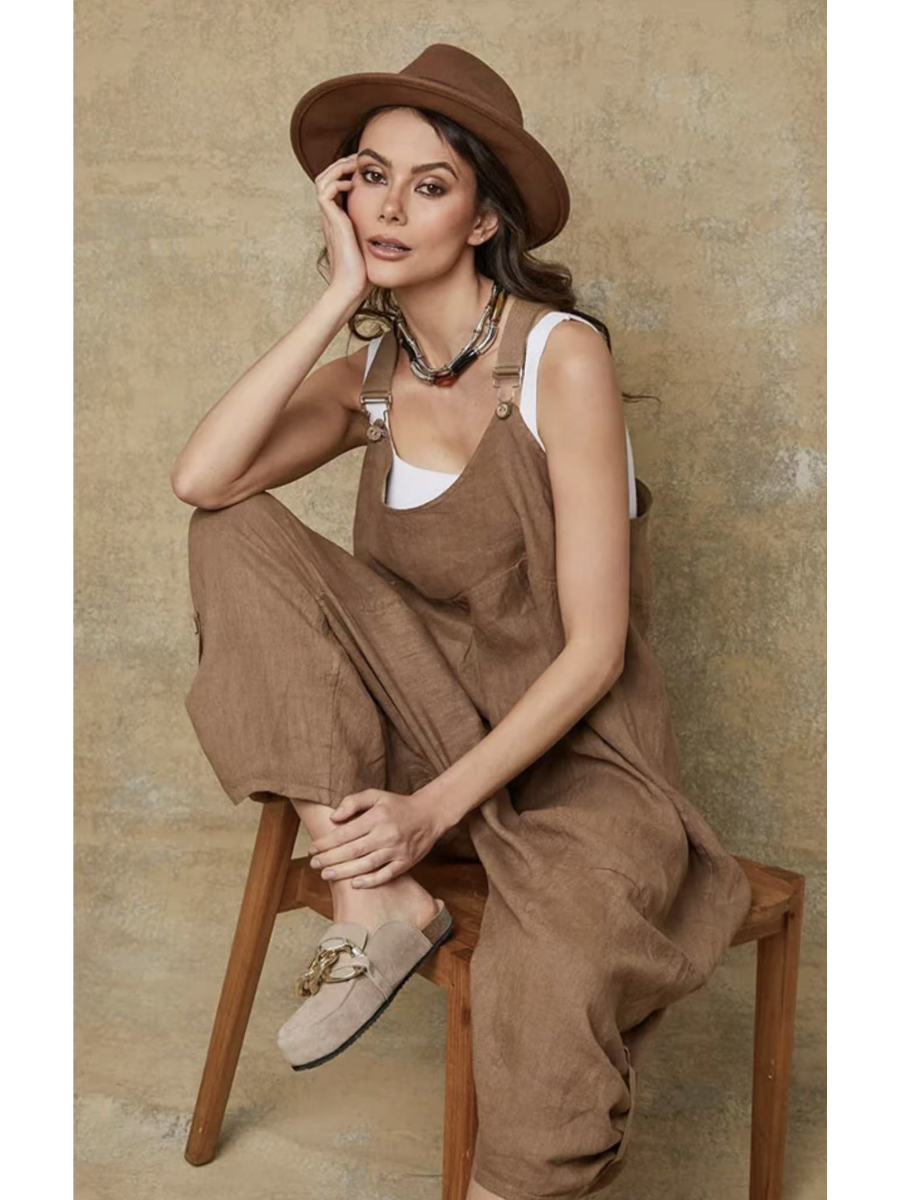 Model in a camel 100% linen jumpsuit with a relaxed fit, adjustable buckle straps, and button details at the ankles, over a white tank top.