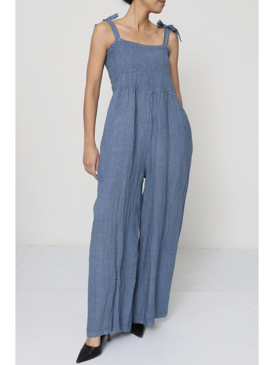 Woman wearing a jeans blue 100% linen jumpsuit with a smocked bodice, tie-shoulder straps, and wide-leg trousers.