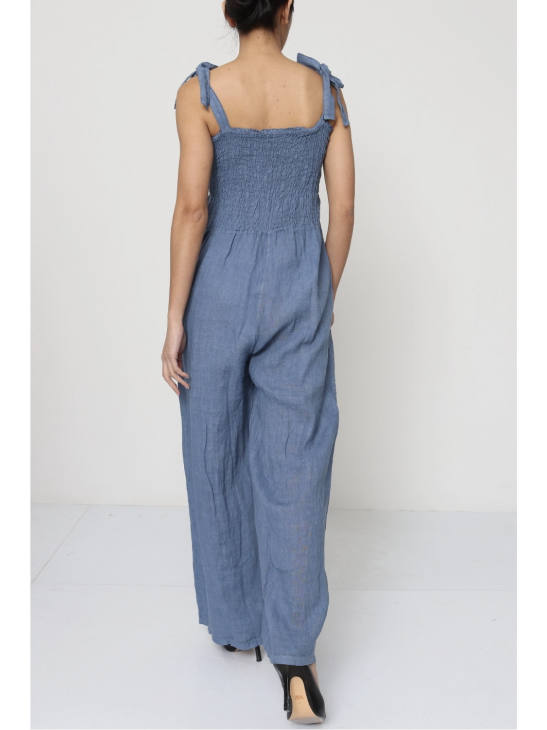 Woman wearing a jeans blue 100% linen jumpsuit with a smocked bodice, tie-shoulder straps, and wide-leg trousers.