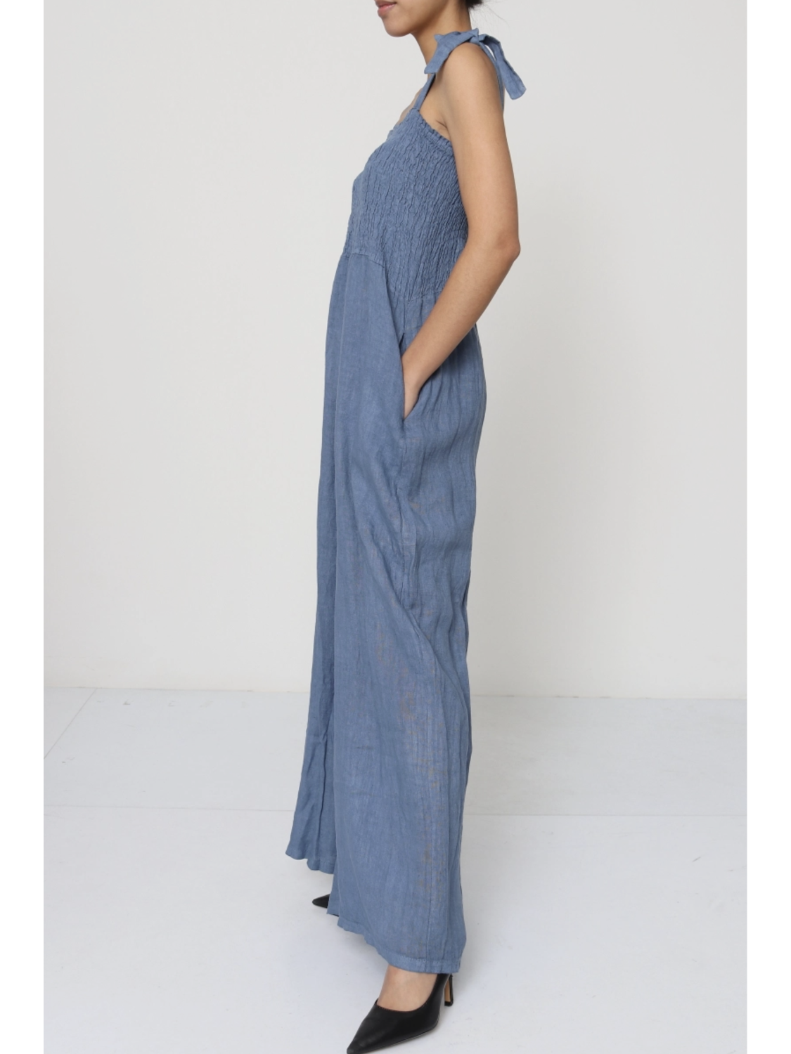 Woman wearing a jeans blue 100% linen jumpsuit with a smocked bodice, tie-shoulder straps, and wide-leg trousers.