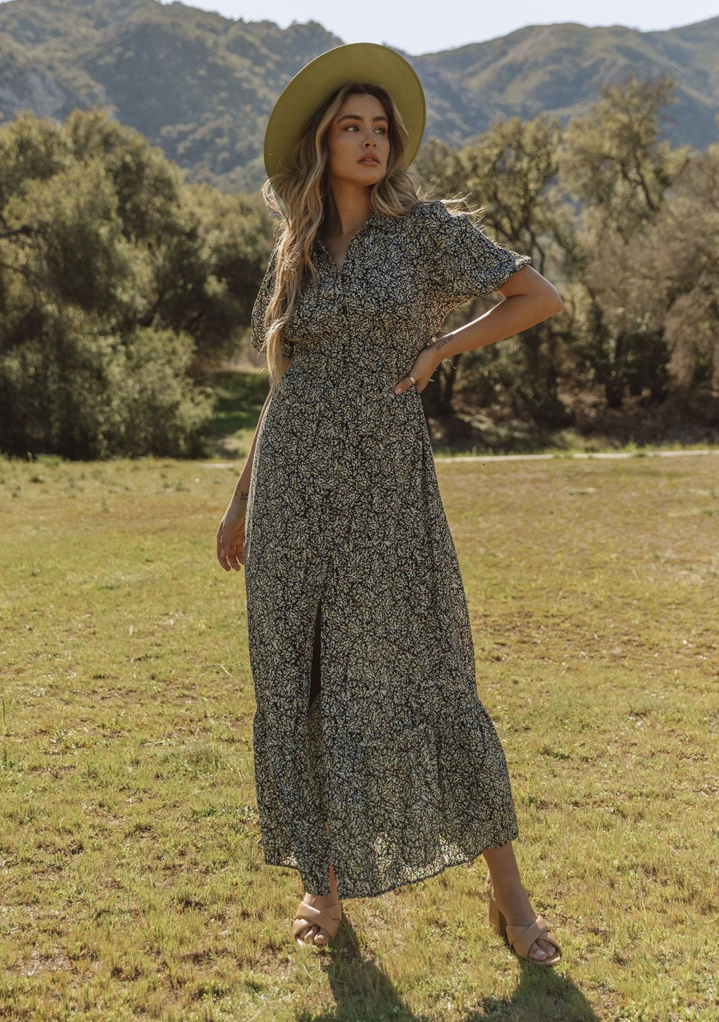 Woman in a floral dress and wide-brimmed hat standing in a field with mountains in the background