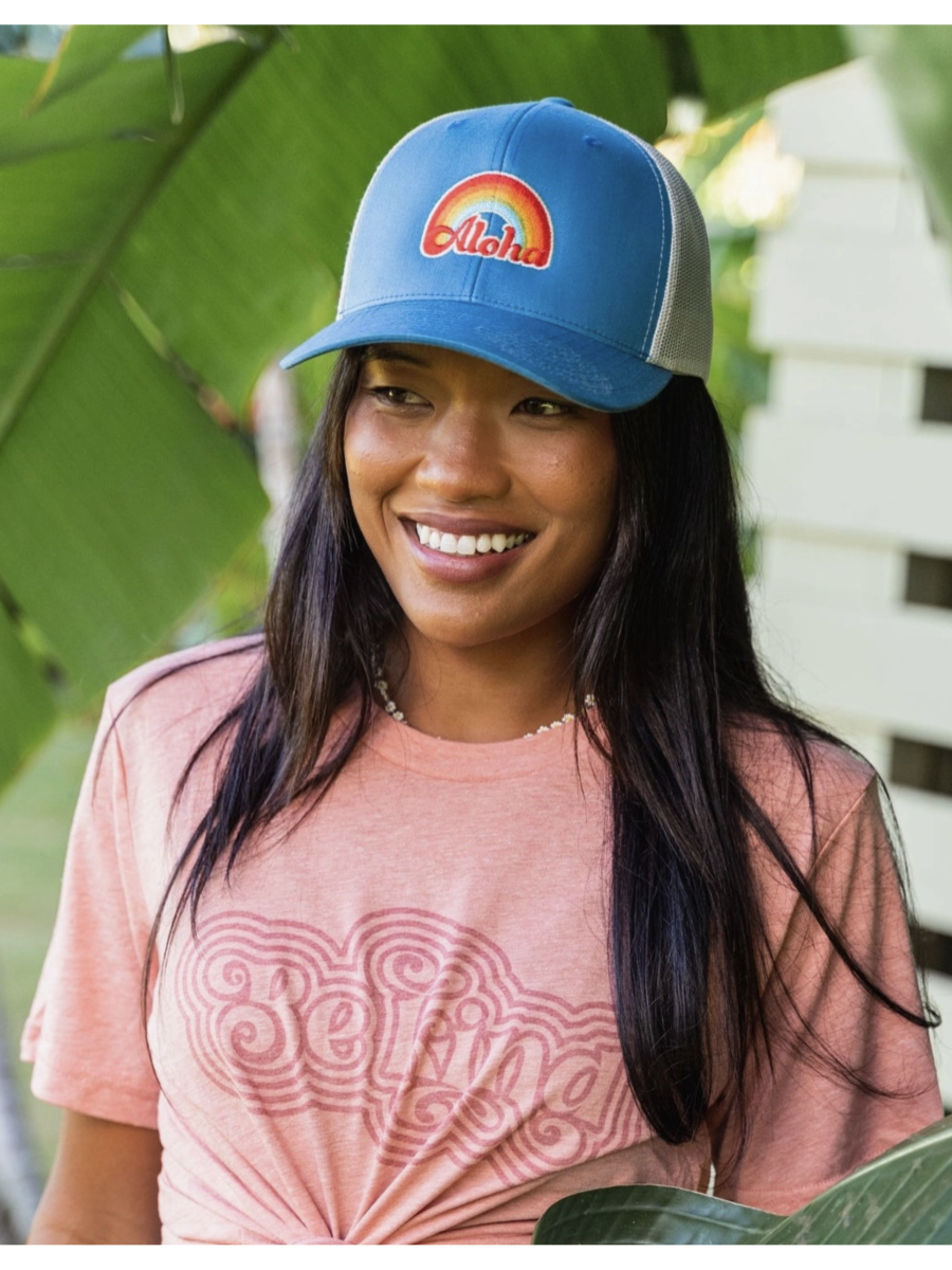 Woman wearing a blue cap with a colorful logo and 'Aloha' text, standing outdoors with greenery in the background.