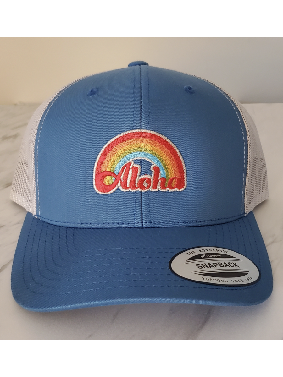 Blue and white trucker hat with a rainbow and 'Aloha' design on a beige background