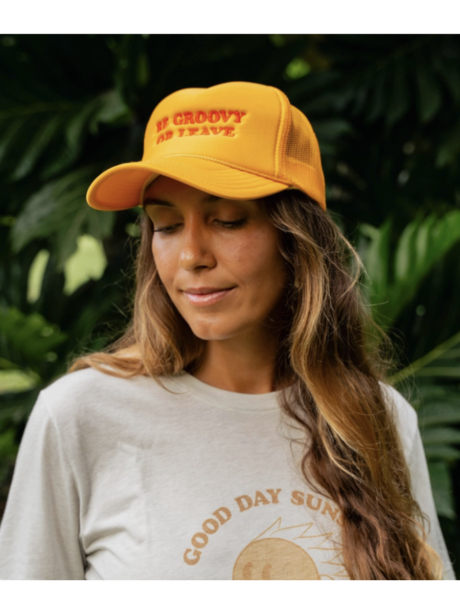 Woman wearing a yellow cap with text 'be groovy or leave' and a 'Good Day Sun' shirt in front of green foliage