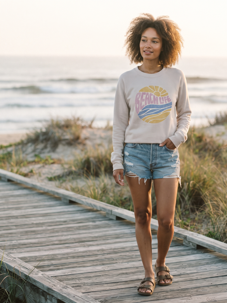A woman stands on a wooden boardwalk by the beach wearing a light beige crewneck sweatshirt with a colorful 'Beach Life' graphic, denim shorts, and sandals.