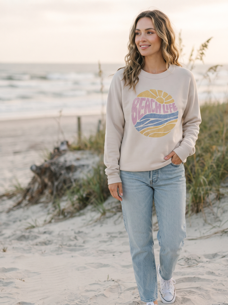 A woman stands on a sandy beach wearing a beige crewneck sweatshirt with a colorful retro circular graphic that reads 'BEACH LIFE,' paired with light blue jeans and white sneakers.