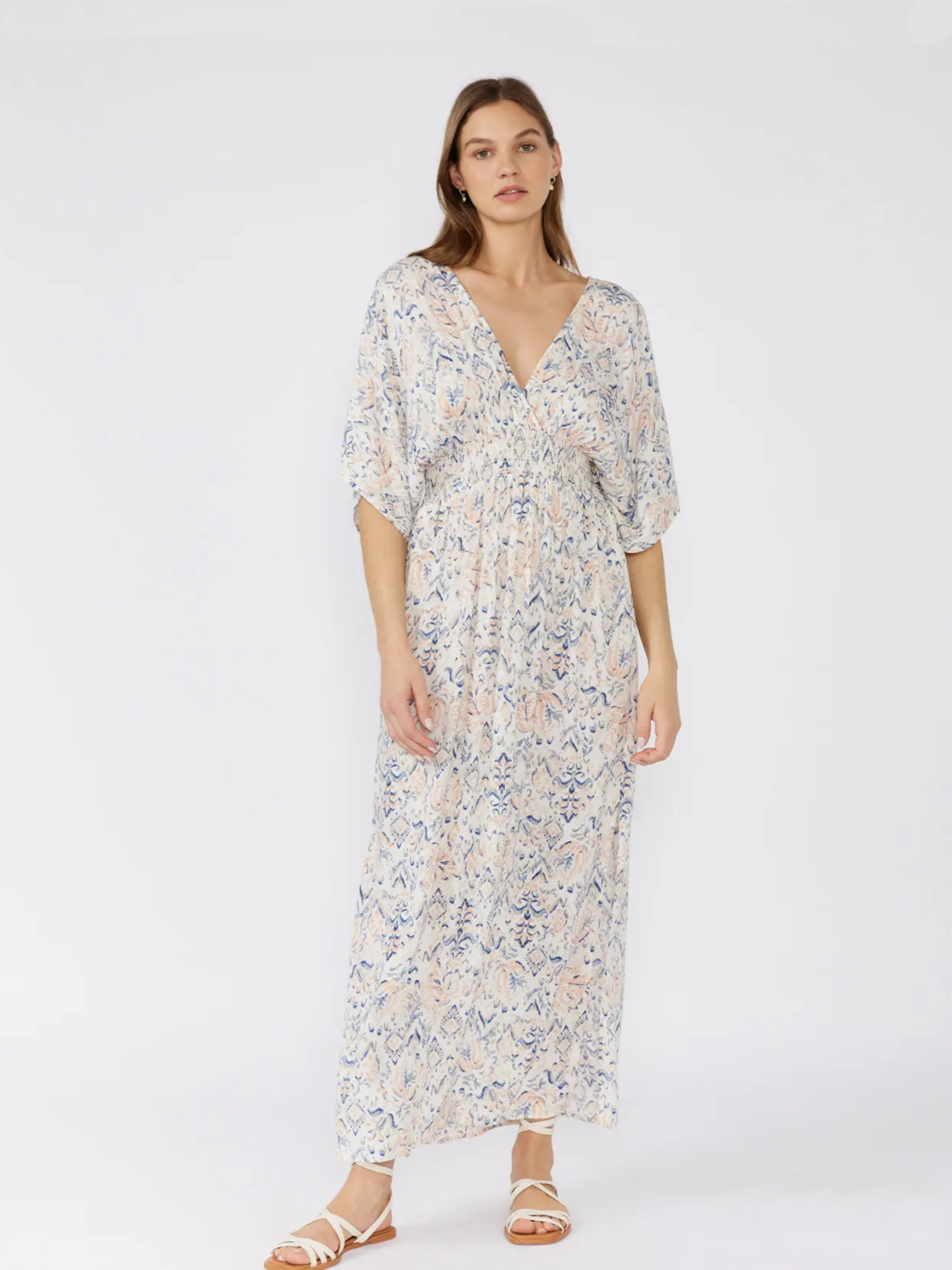 Woman in a white bohemian maxi dress with a blue and peach paisley print, kimono sleeves, and a smocked waist, standing against a white background.