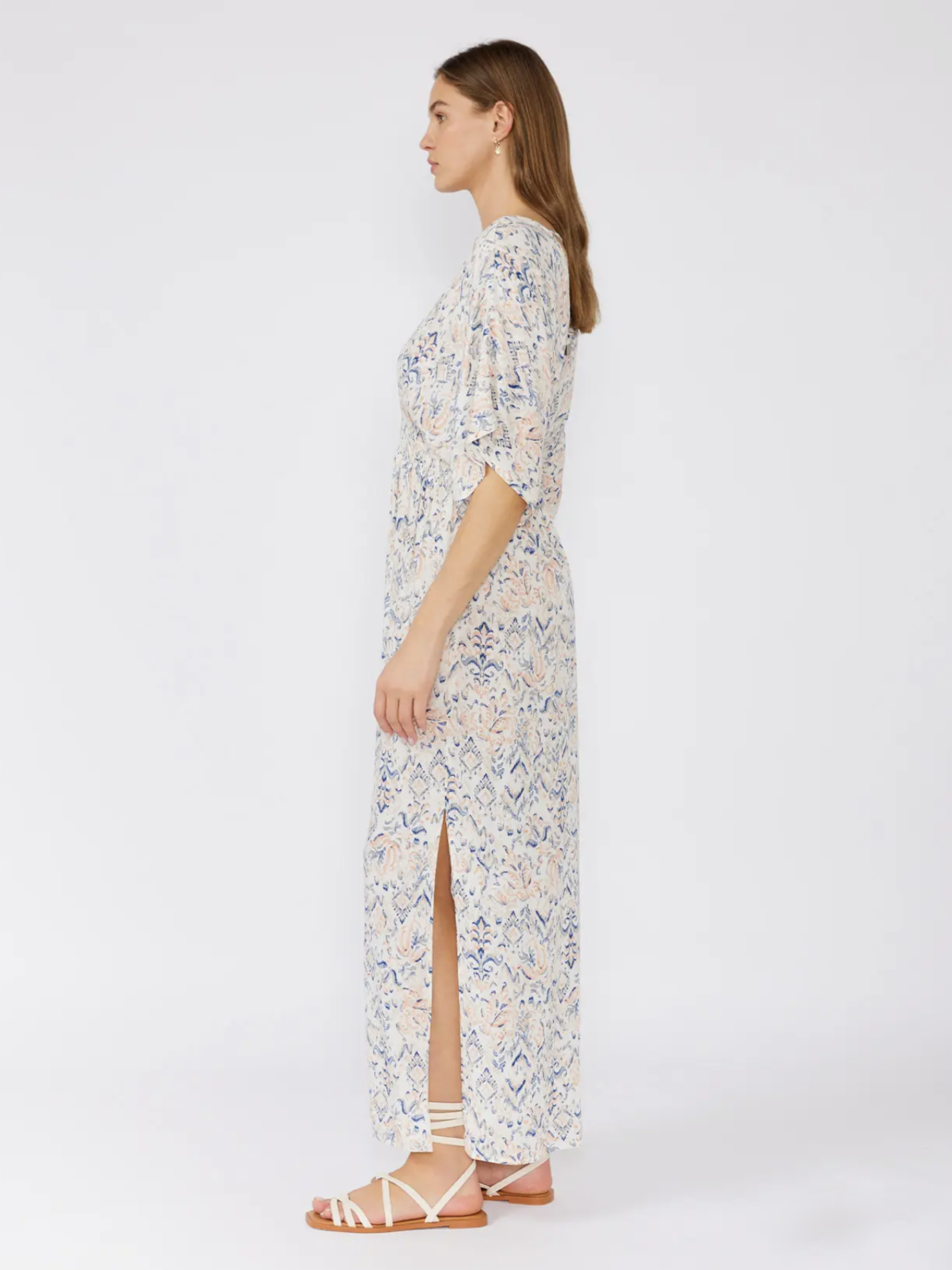 Woman in a white bohemian maxi dress with a blue and peach paisley print, kimono sleeves, and a smocked waist, standing against a white background.