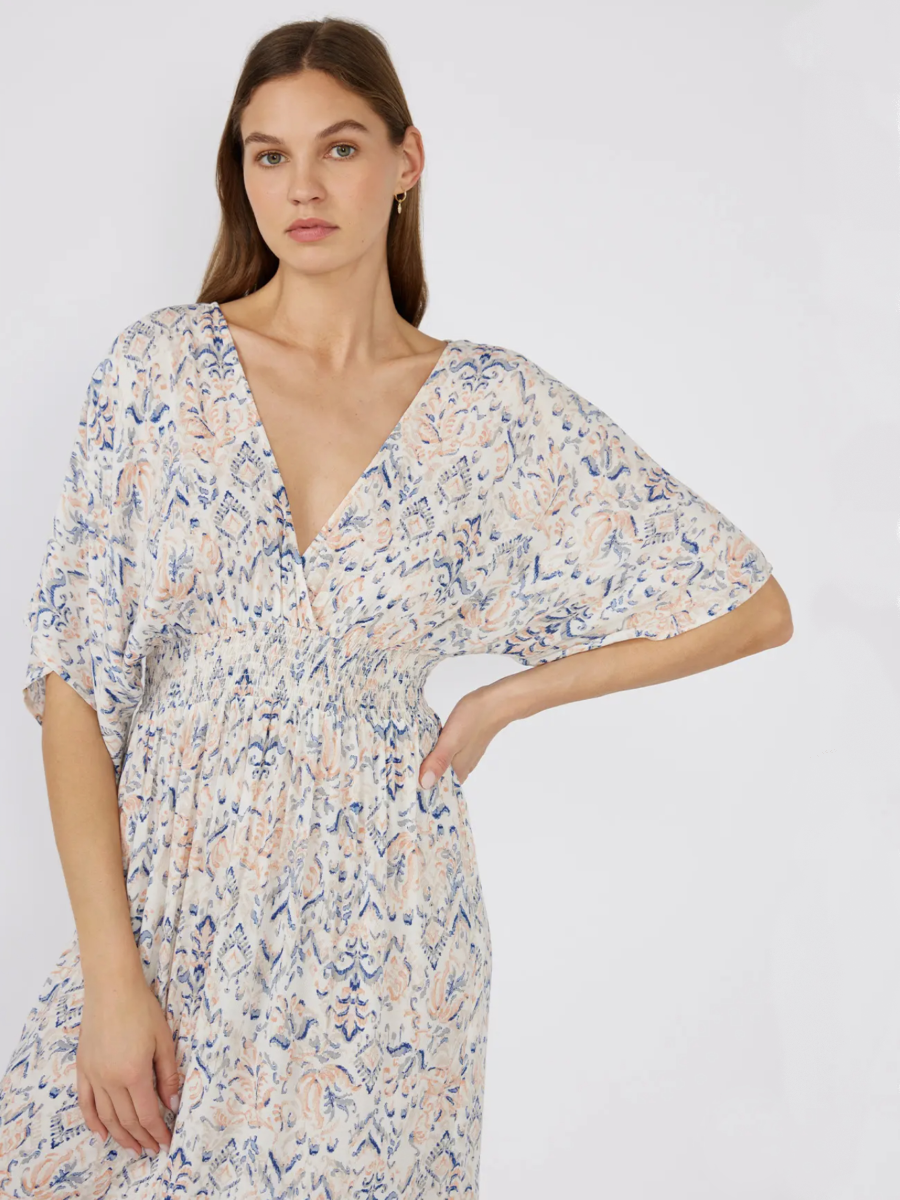 Woman in a white bohemian maxi dress with a blue and peach paisley print, kimono sleeves, and a smocked waist, standing against a white background.