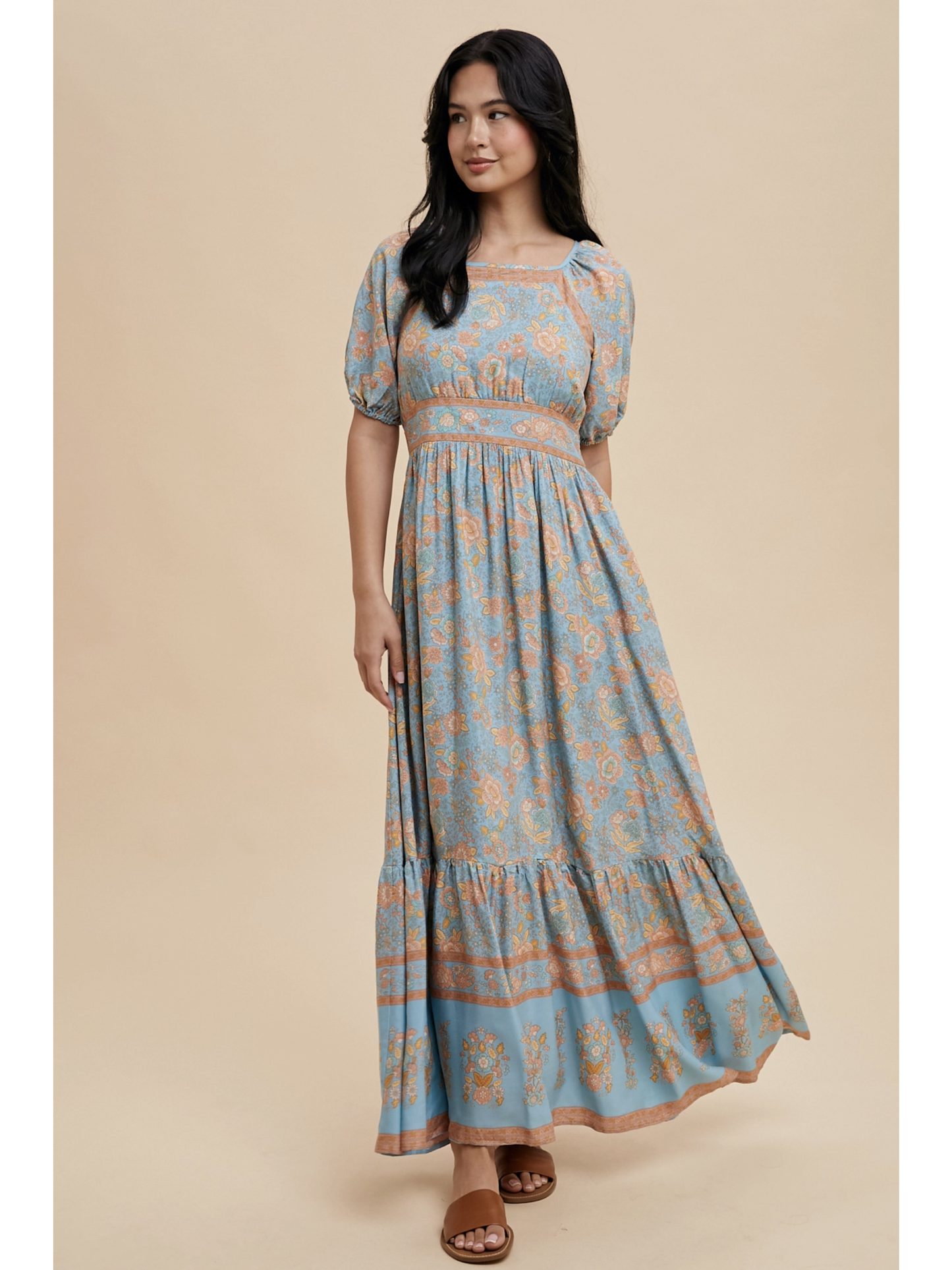A woman wearing a dusty blue maxi dress with white botanical border prints, lace trim, standing against a soft-focus background.