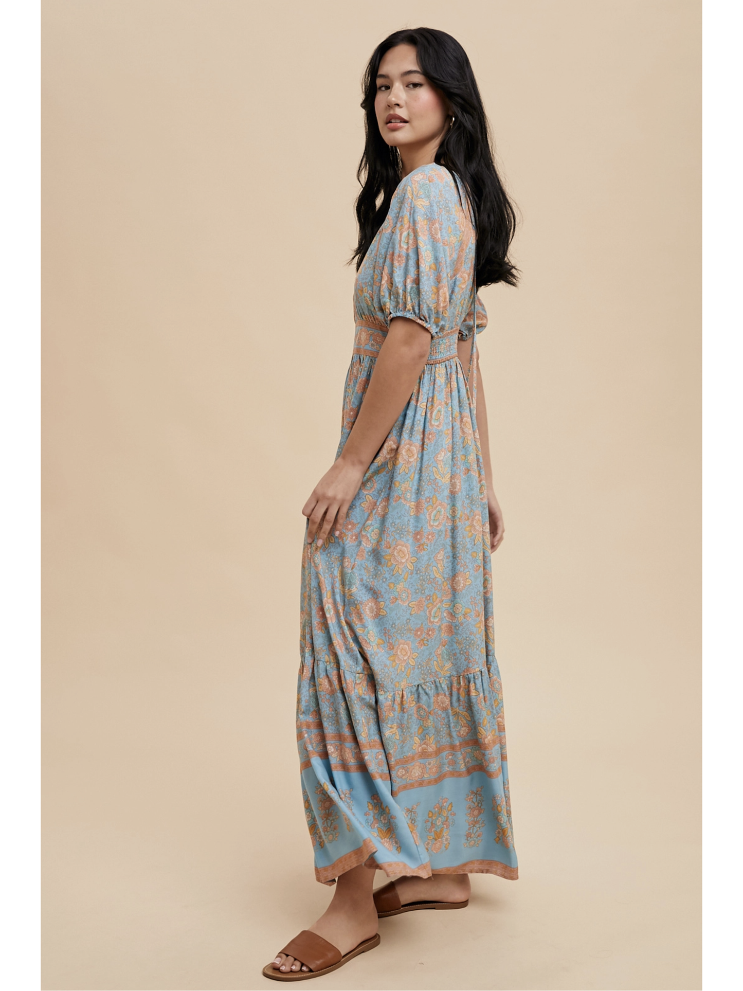 A woman wearing a dusty blue maxi dress with white botanical border prints, lace trim, standing against a soft-focus background.