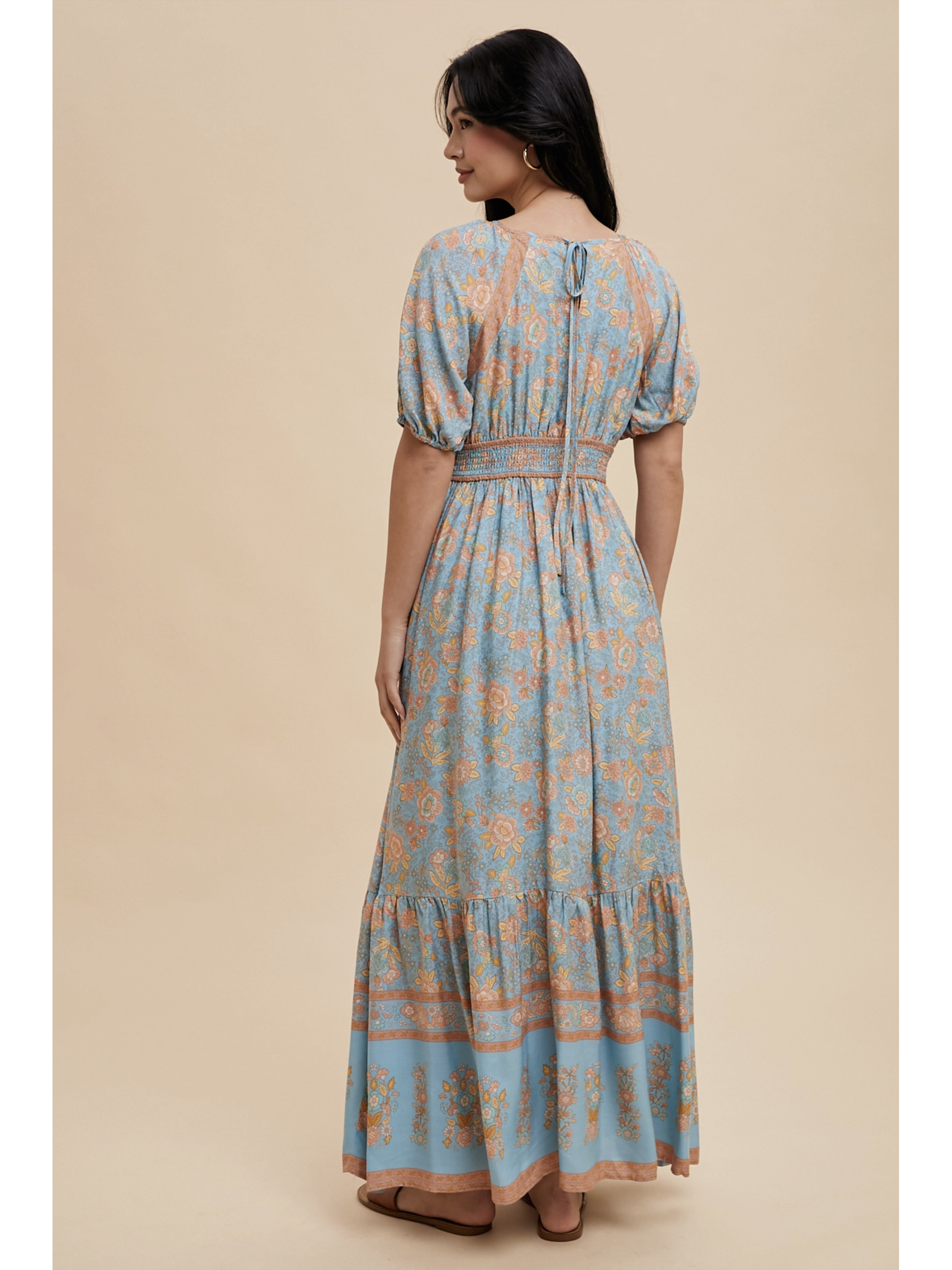 A woman wearing a dusty blue maxi dress with white botanical border prints, lace trim, standing against a soft-focus background.