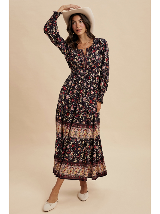Woman in a midnight black tiered maxi dress with red floral patterns and a wide ornamental border at the hem, wearing a light-colored felt hat.