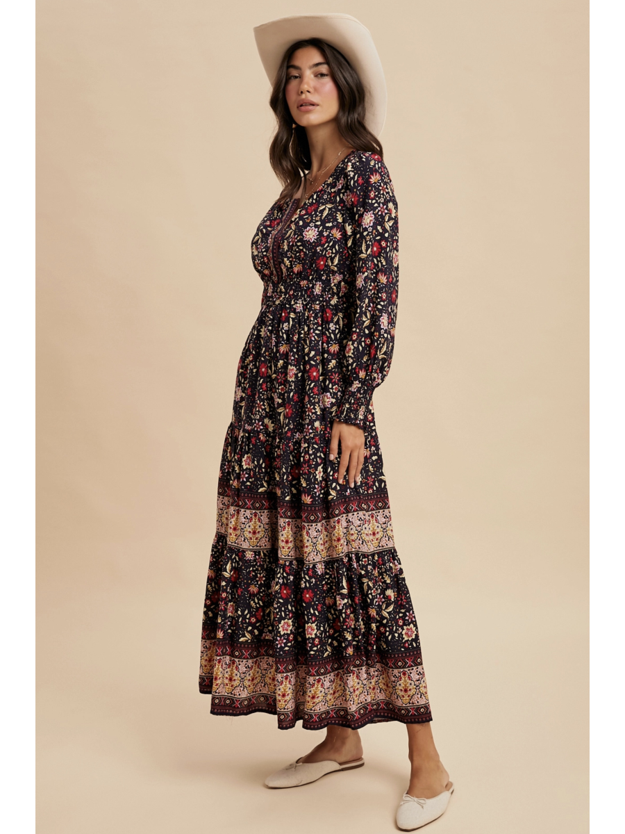 Woman in a midnight black tiered maxi dress with red floral patterns and a wide ornamental border at the hem, wearing a light-colored felt hat.
