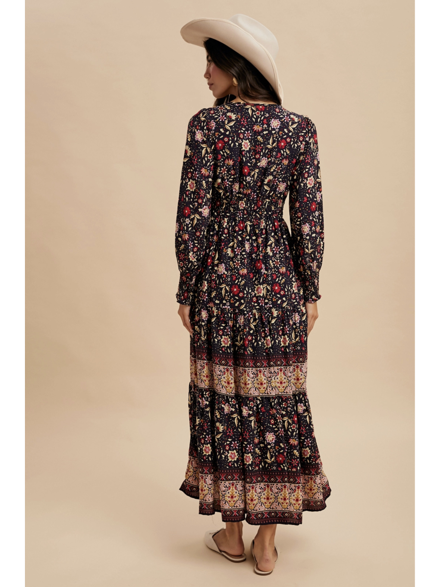 Woman in a midnight black tiered maxi dress with red floral patterns and a wide ornamental border at the hem, wearing a light-colored felt hat.
