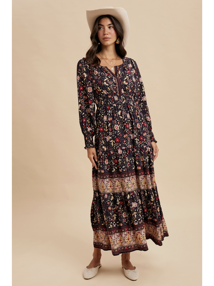 Woman in a midnight black tiered maxi dress with red floral patterns and a wide ornamental border at the hem, wearing a light-colored felt hat.