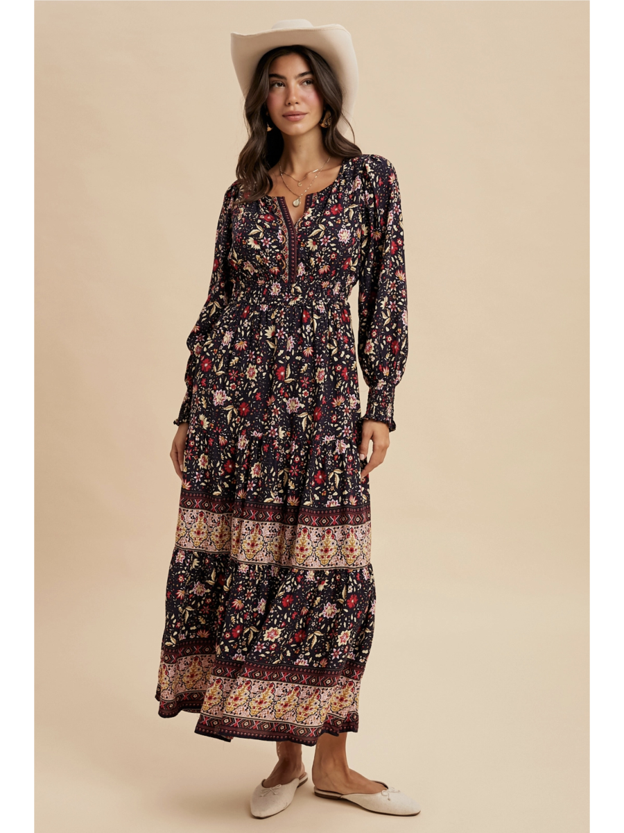 Woman in a midnight black tiered maxi dress with red floral patterns and a wide ornamental border at the hem, wearing a light-colored felt hat.