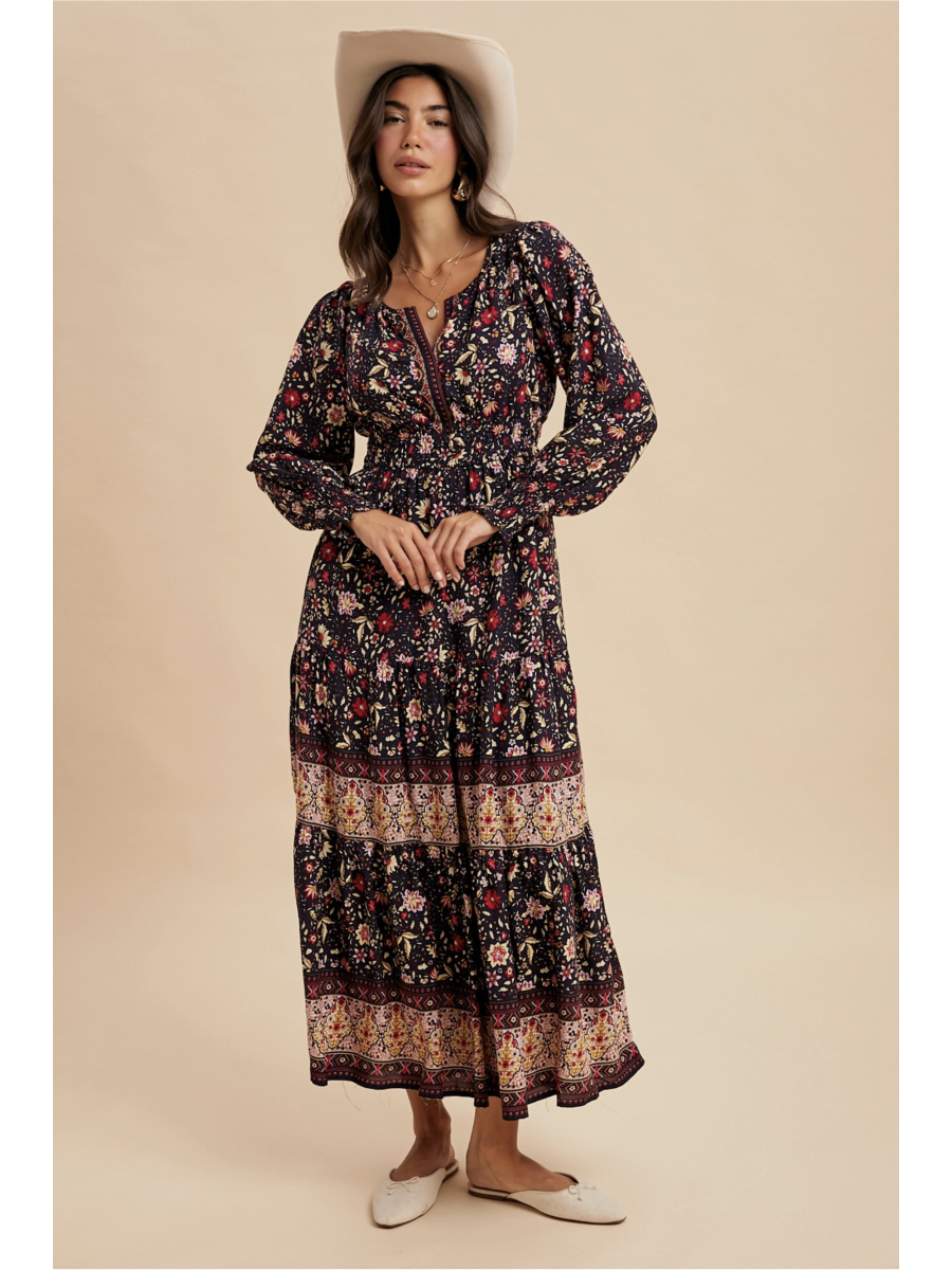 Woman in a midnight black tiered maxi dress with red floral patterns and a wide ornamental border at the hem, wearing a light-colored felt hat.