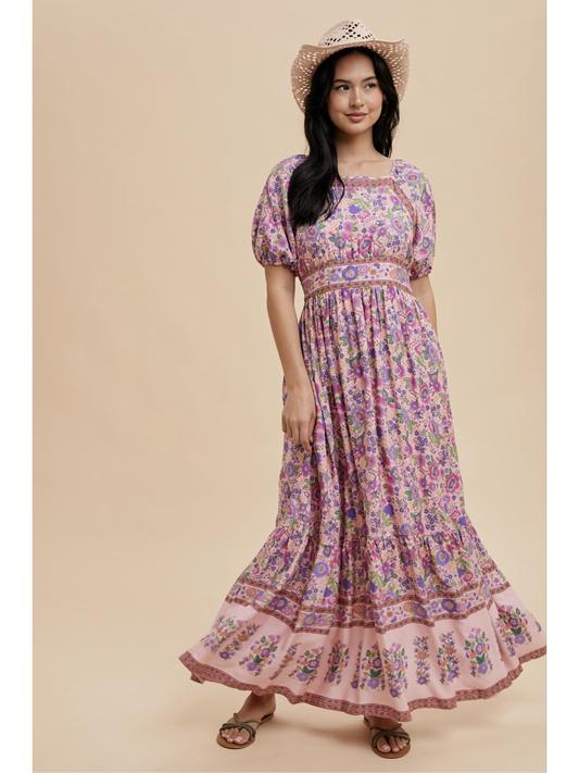 A woman wearing a pink fuschia maxi dress with white botanical border prints, lace trim, standing against a soft-focus background.