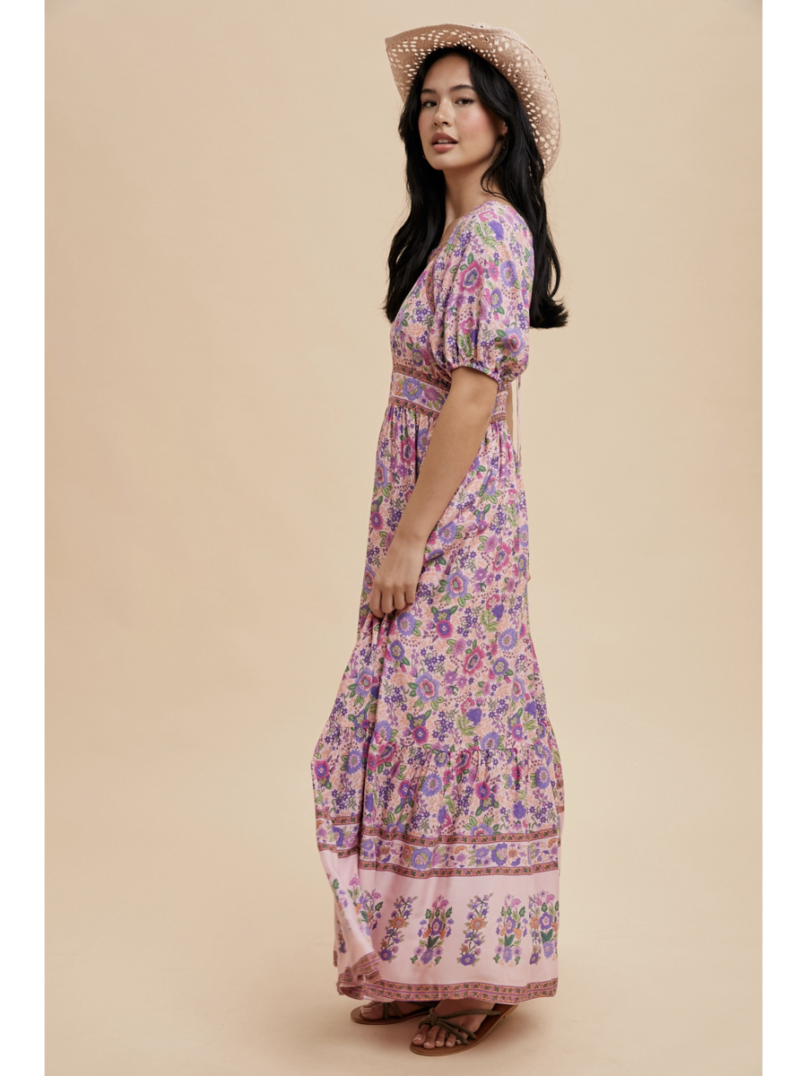 A woman wearing a pink fuschia maxi dress with white botanical border prints, lace trim, standing against a soft-focus background.