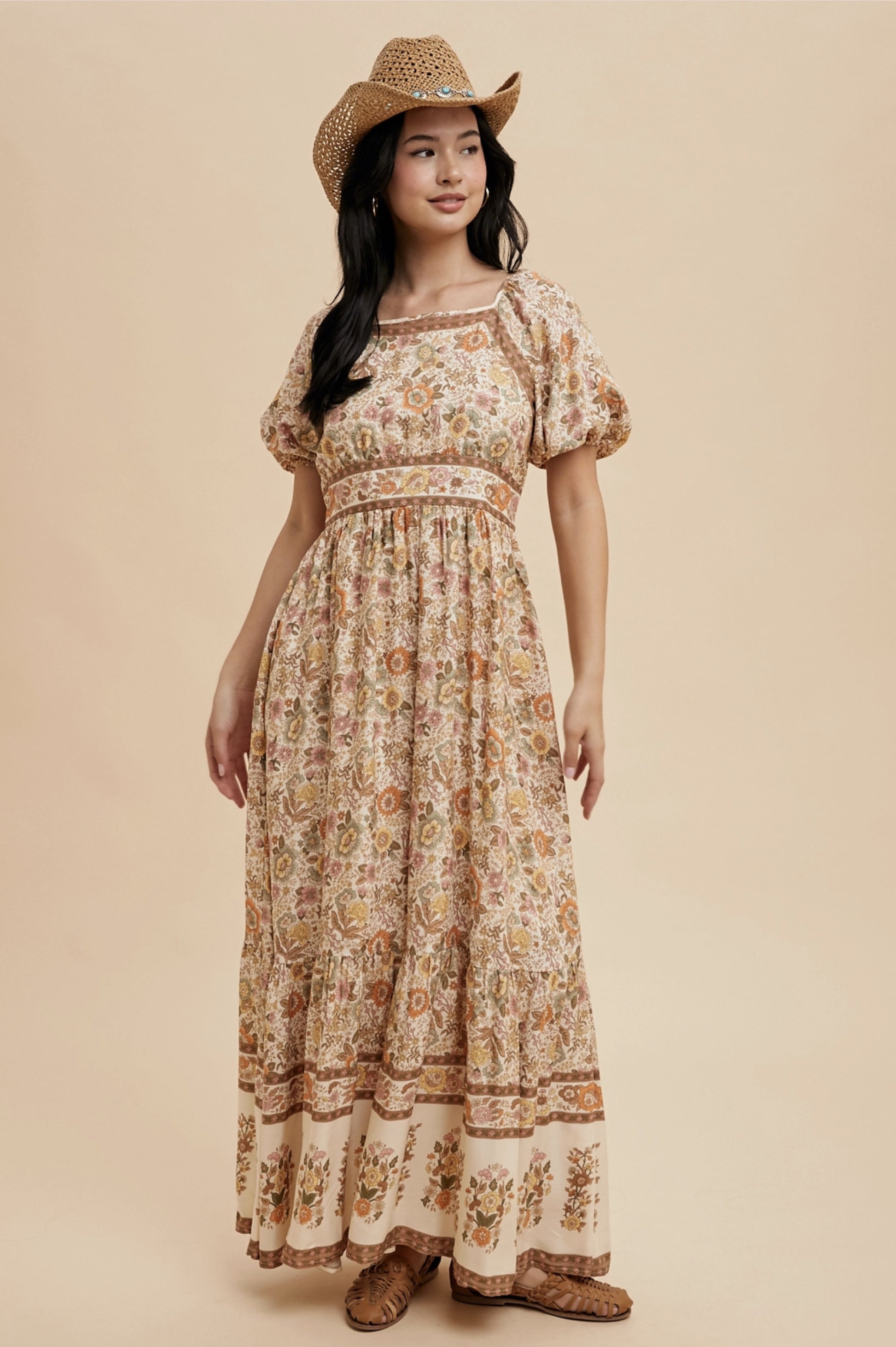 A long, floral print dress with a square neckline and short puff sleeves. The dress features a cream-colored background with a repeating pattern of flowers in shades of orange, yellow, and brown. A straw hat completes the look.