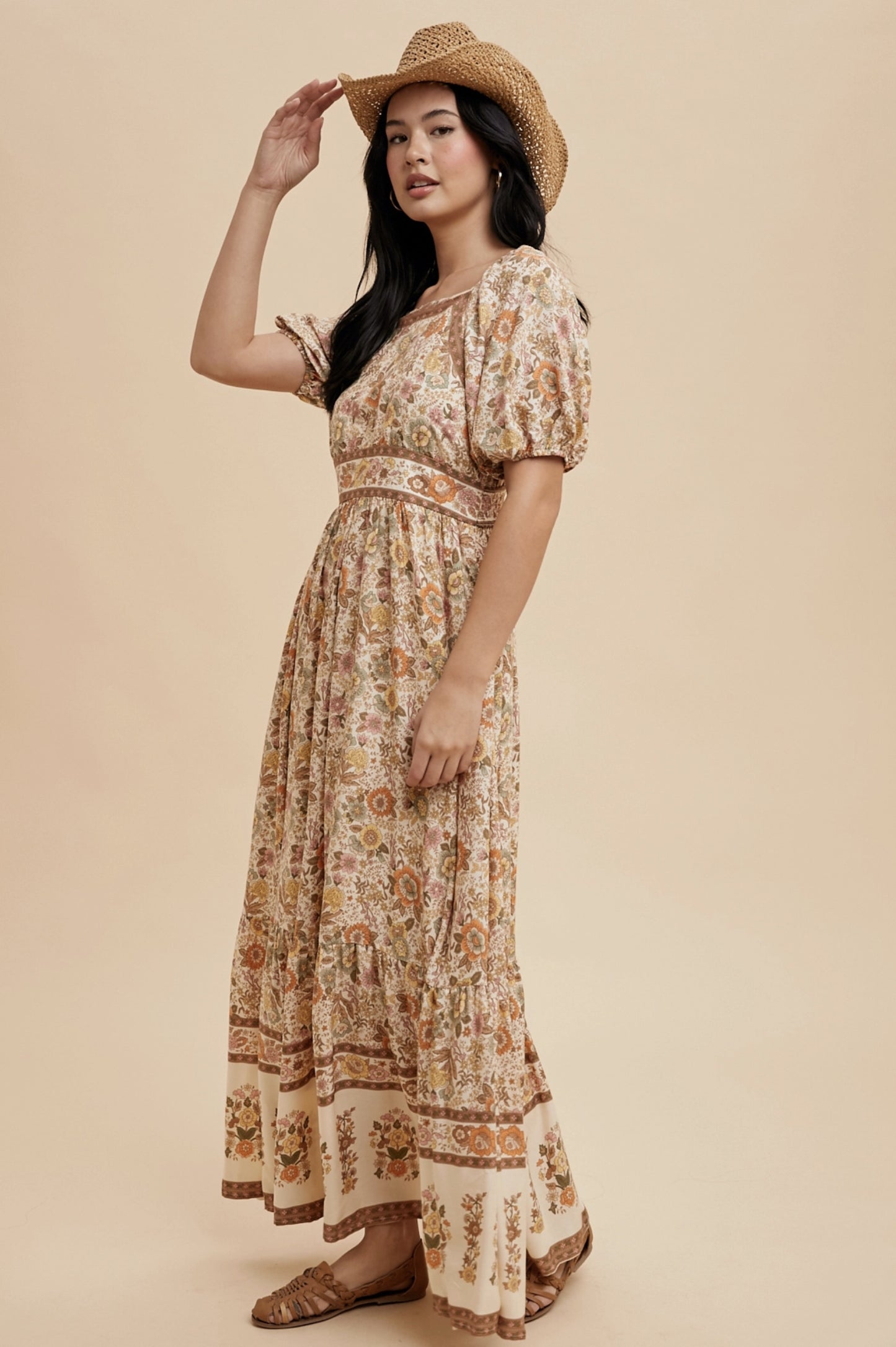 A long, floral print dress with a square neckline and short puff sleeves. The dress features a cream-colored background with a repeating pattern of flowers in shades of orange, yellow, and brown. A straw hat completes the look.