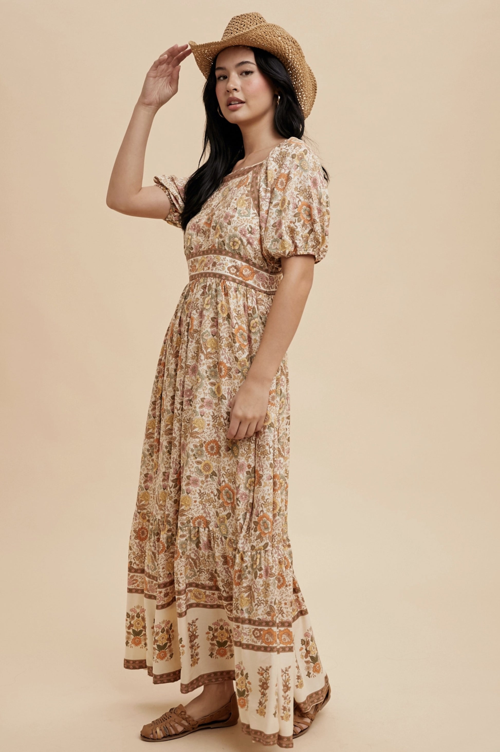 A long, floral print dress with a square neckline and short puff sleeves. The dress features a cream-colored background with a repeating pattern of flowers in shades of orange, yellow, and brown. A straw hat completes the look.