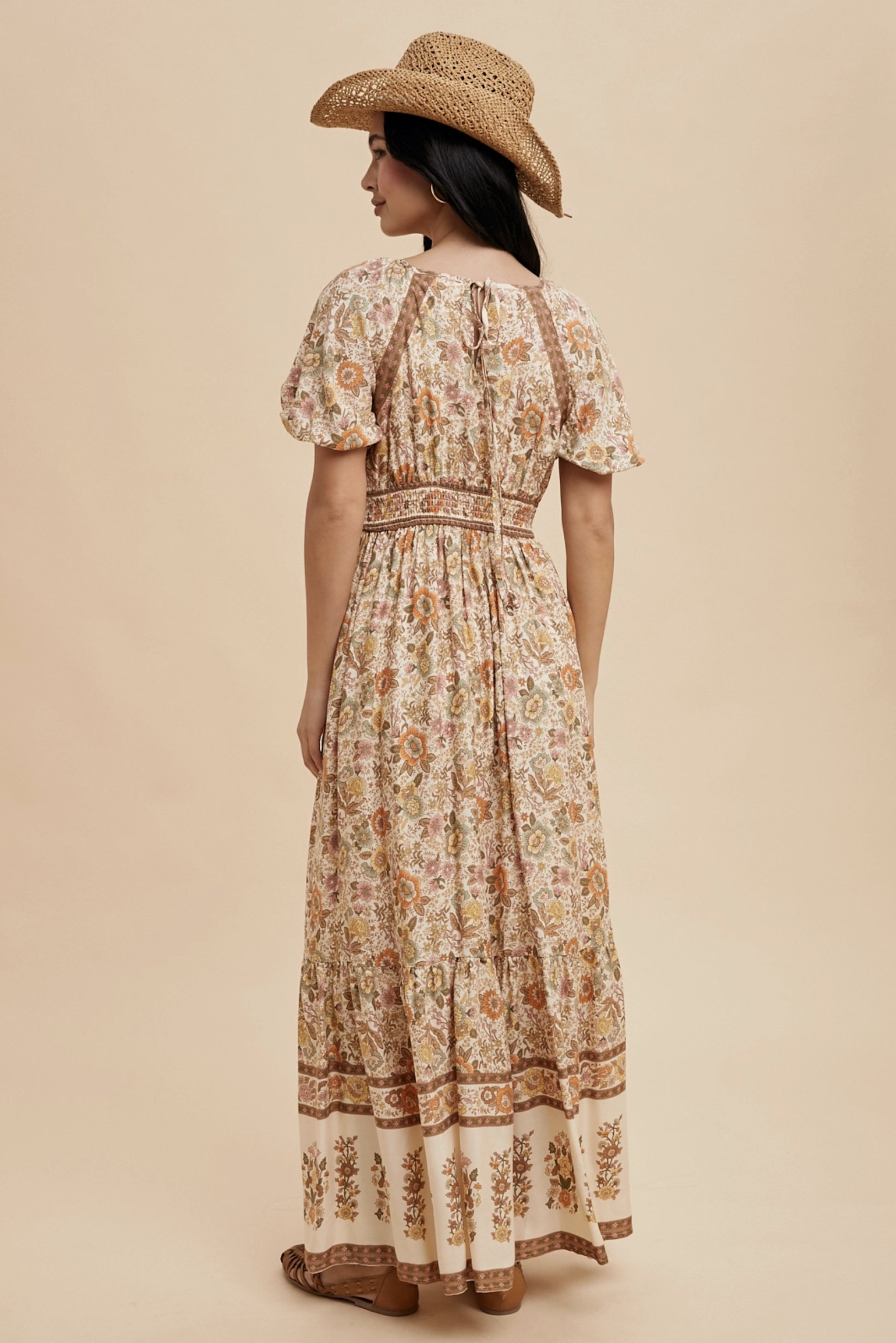 A long, floral print dress with a square neckline and short puff sleeves. The dress features a cream-colored background with a repeating pattern of flowers in shades of orange, yellow, and brown. A straw hat completes the look.