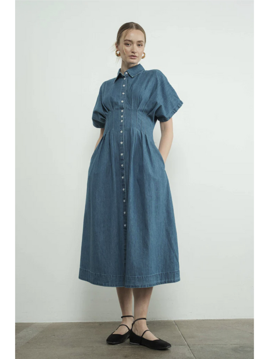 A woman wearing a denim blue midi shirt dress with a button-front bodice, pointed collar, matching waist belt, and a flowy pleated skirt.