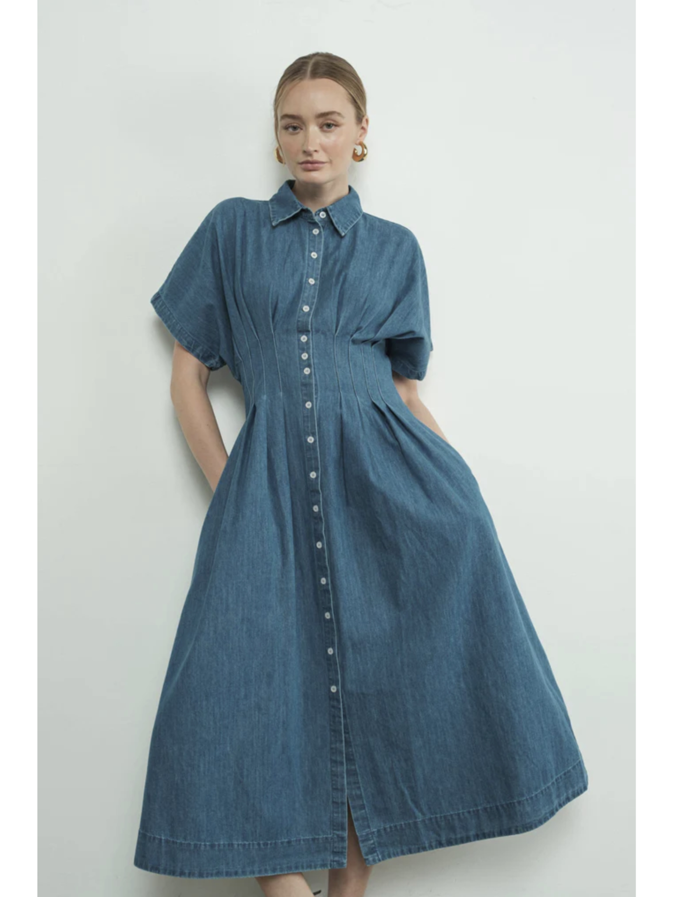 A woman wearing a denim blue midi shirt dress with a button-front bodice, pointed collar, matching waist belt, and a flowy pleated skirt.