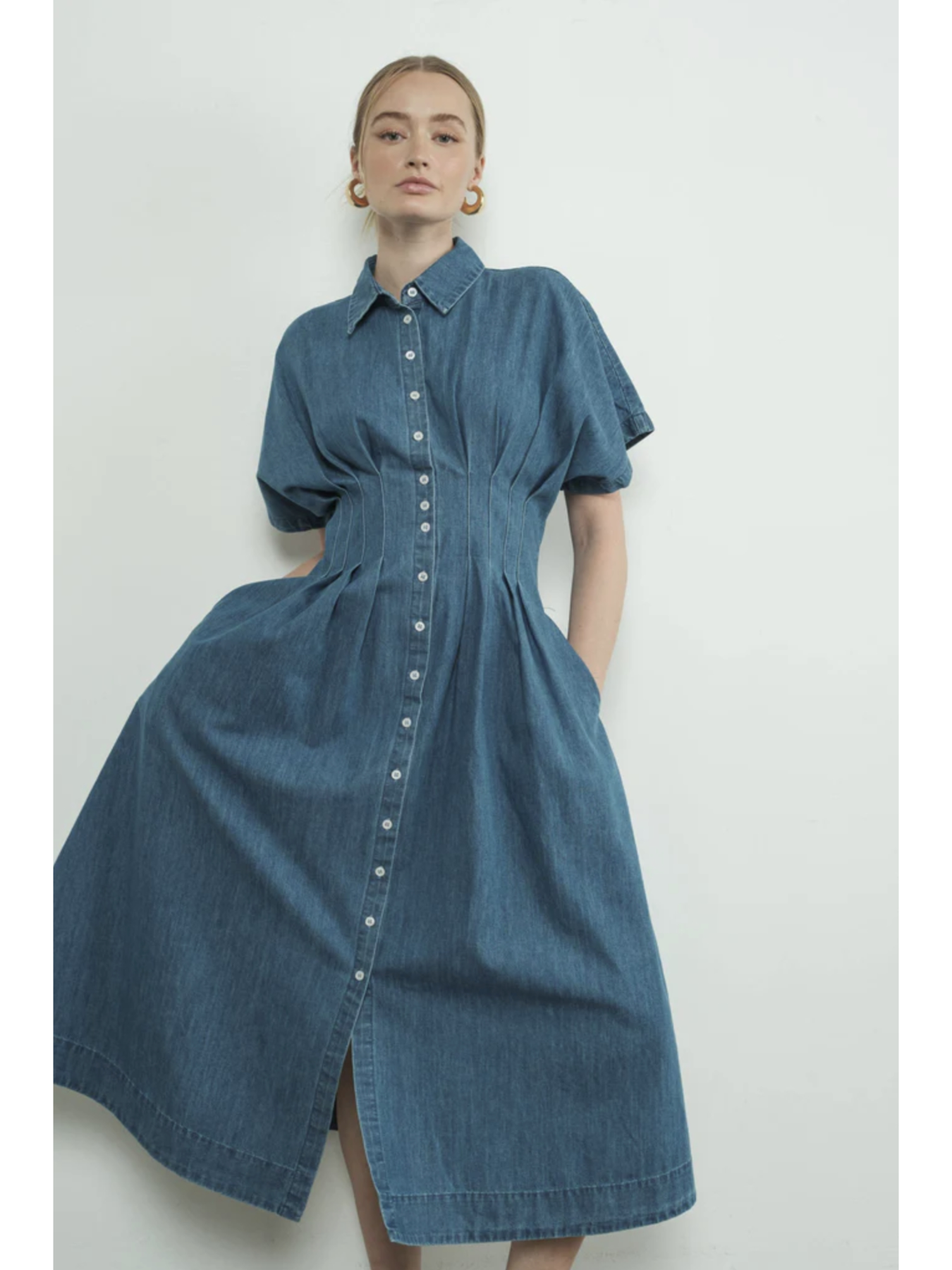 A woman wearing a denim blue midi shirt dress with a button-front bodice, pointed collar, matching waist belt, and a flowy pleated skirt.