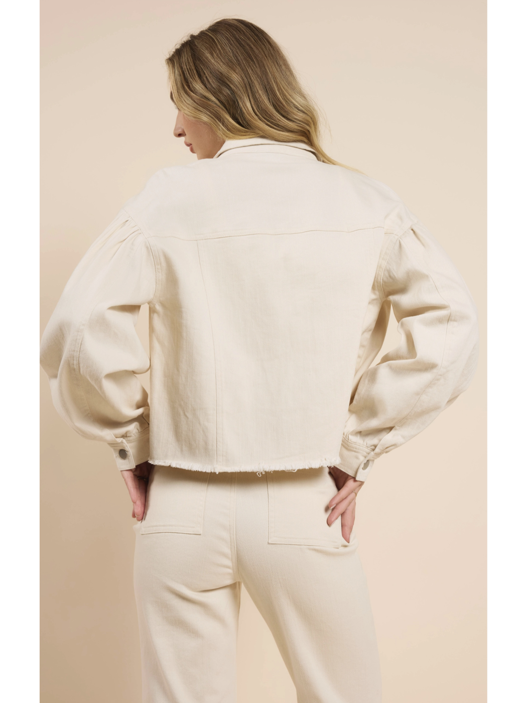 A woman wearing a cream-colored cropped denim jacket with dramatic puff sleeves, a raw frayed hem, and matching pants against a neutral background.