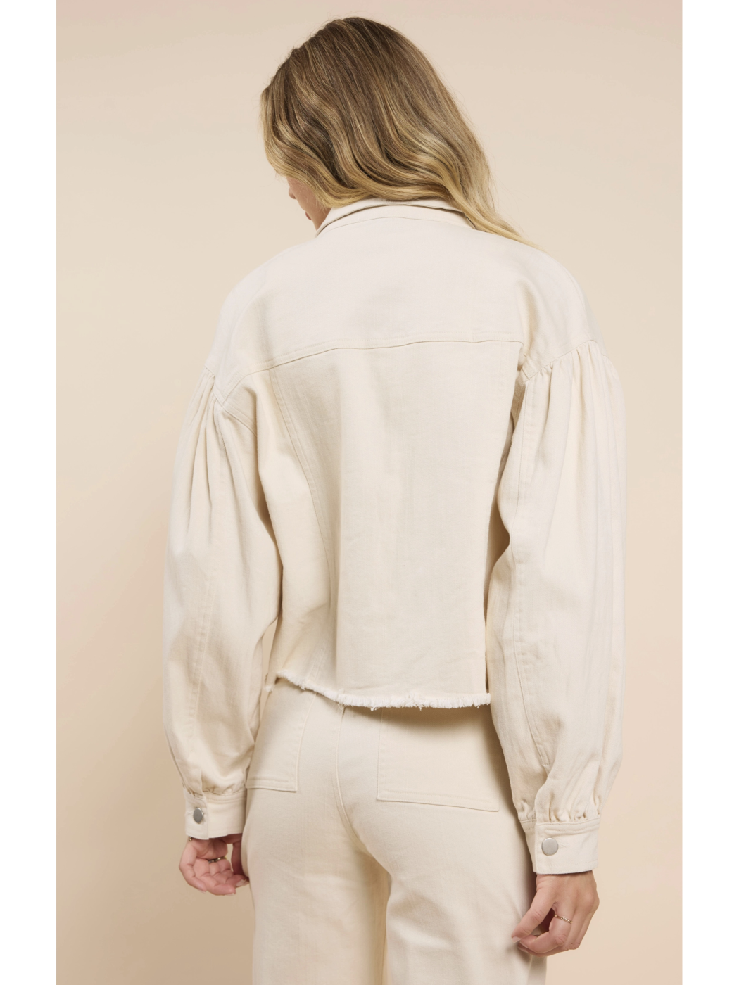 A woman wearing a cream-colored cropped denim jacket with dramatic puff sleeves, a raw frayed hem, and matching pants against a neutral background.