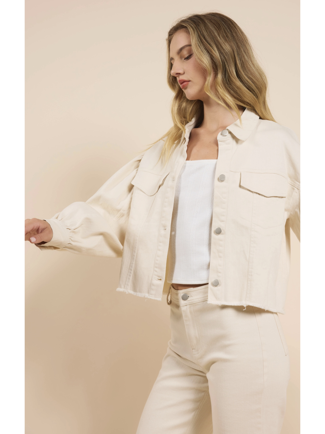 A woman wearing a cream-colored cropped denim jacket with dramatic puff sleeves, a raw frayed hem, and matching pants against a neutral background.