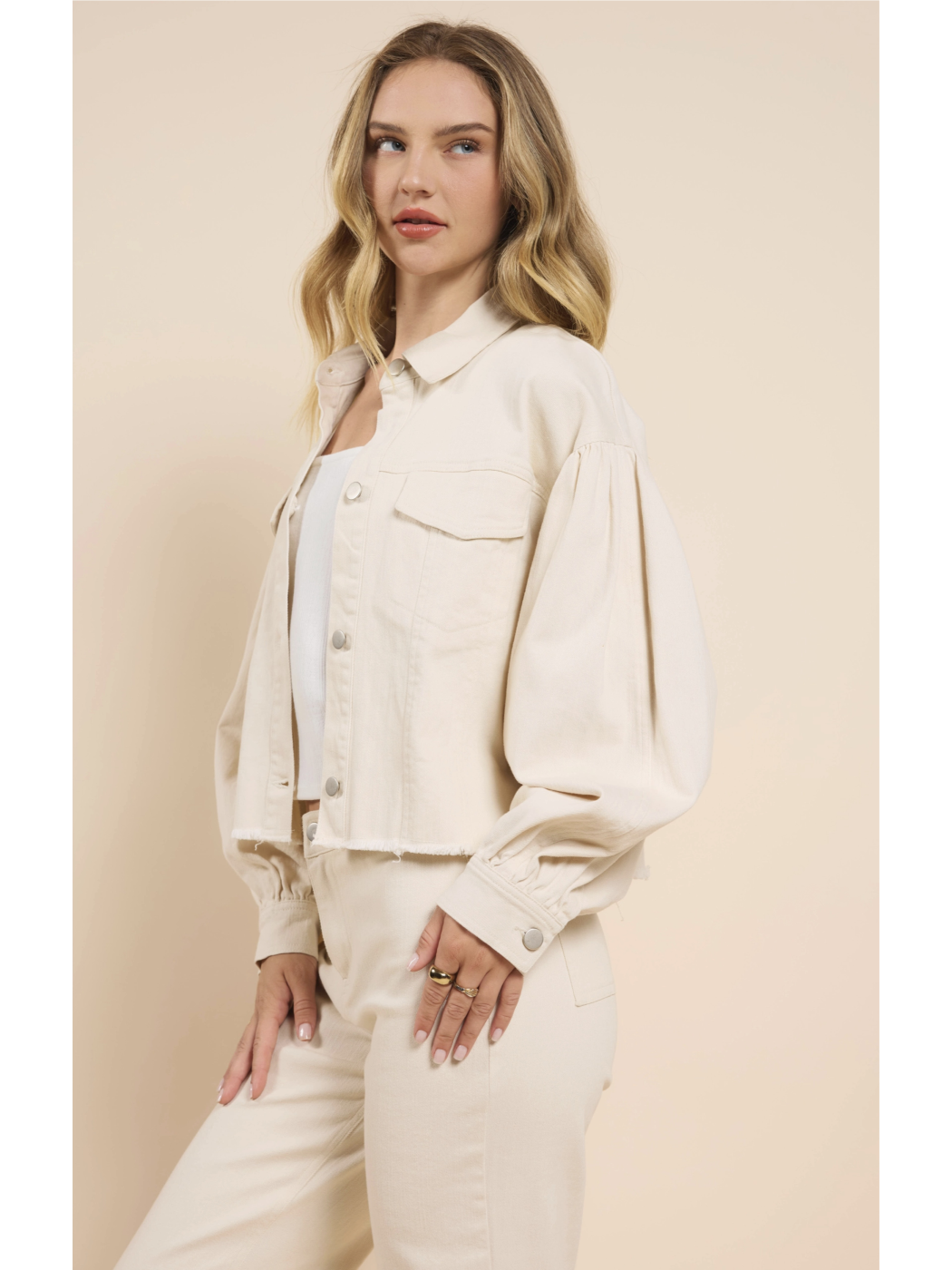 A woman wearing a cream-colored cropped denim jacket with dramatic puff sleeves, a raw frayed hem, and matching pants against a neutral background.
