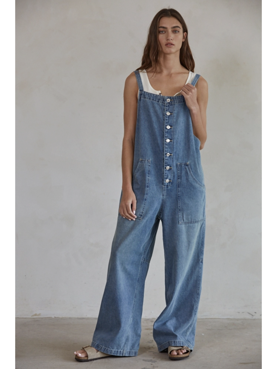 Model in blue denim wide-leg overalls with a square neck and button-down front, styled over a white t-shirt against a plain background.
