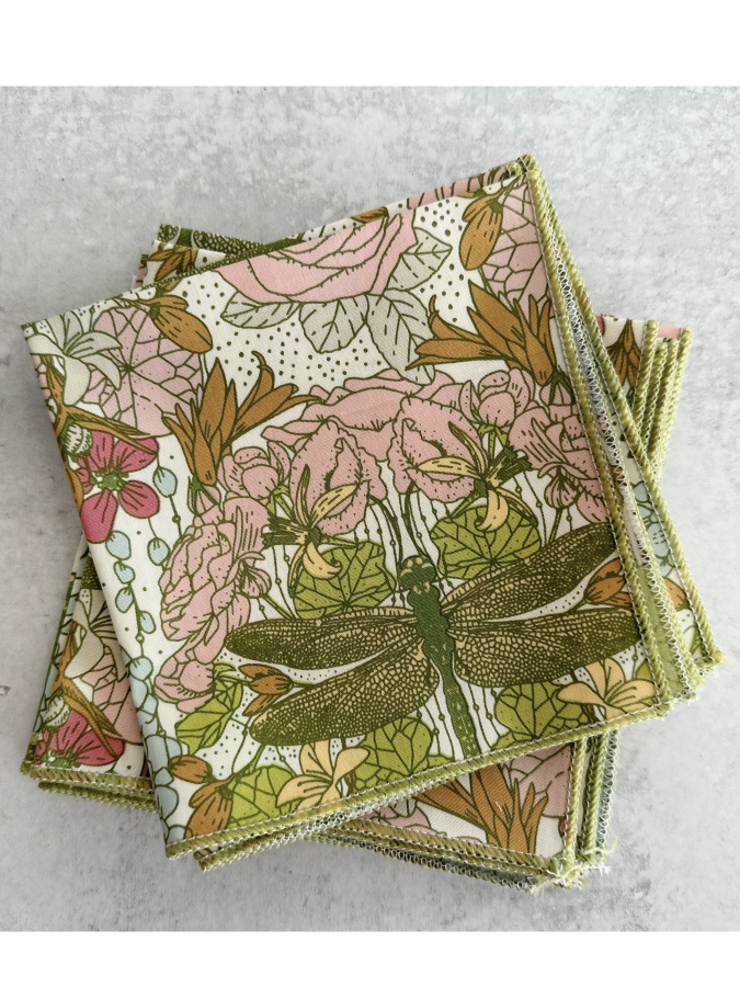 Set of four white cloth napkins with a green dragonfly and pink rose watercolor pattern, folded on a light gray surface.
