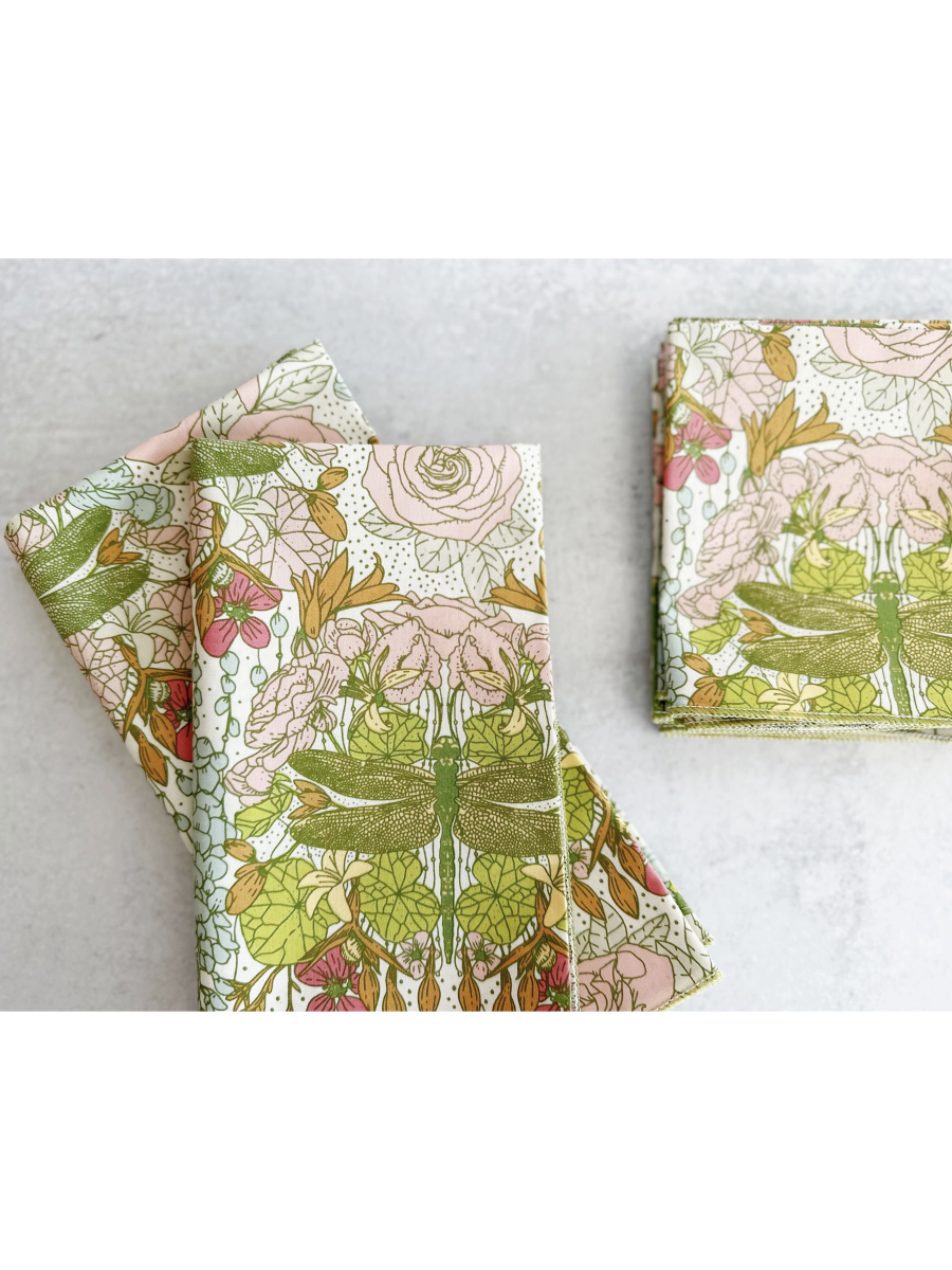 Set of four white cloth napkins with a green dragonfly and pink rose watercolor pattern, folded on a light gray surface.