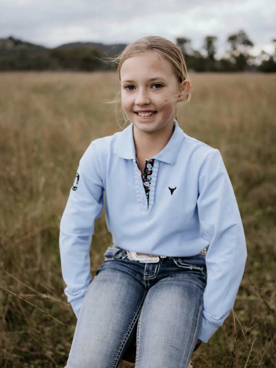 Young girl sitting in a field wearing a light blue sweater with a logo.